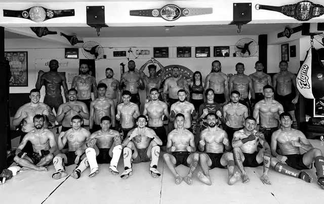 Cerca de trinta atletas do CT Sombra Team posam no tatame da academia, com cinturões de campeonato pendurados na parede acima deles.